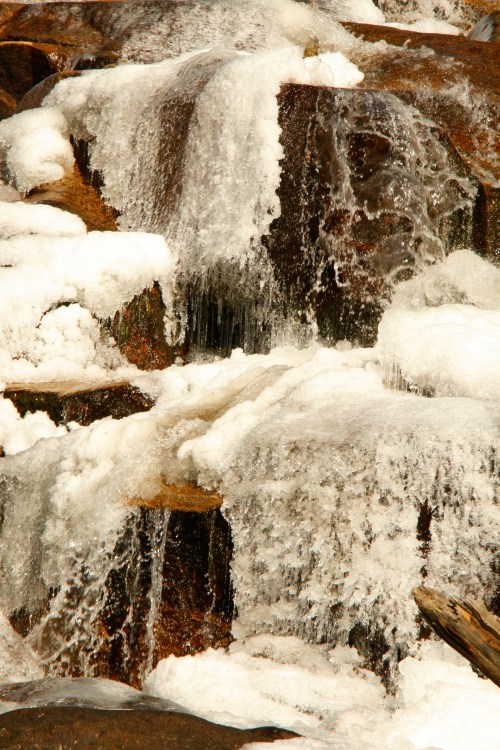 A close-up of a frozen waterfall cascading over rocky surfaces, with ice and snow covering the stones.