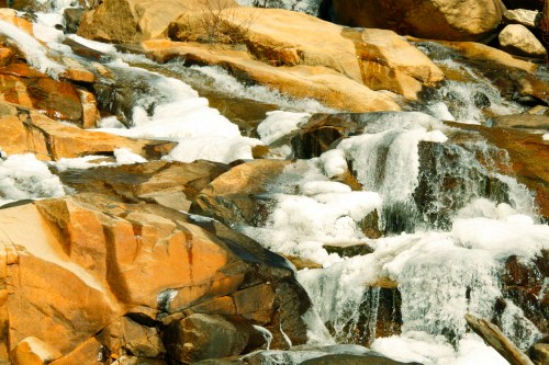 A close-up of a rocky stream with flowing water and frothy white bubbles over smooth stones.