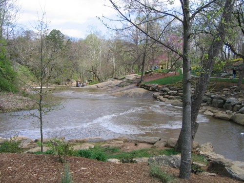 A serene river flows through a wooded area, surrounded by rocks and trees, with people in the distance enjoying the scenery.