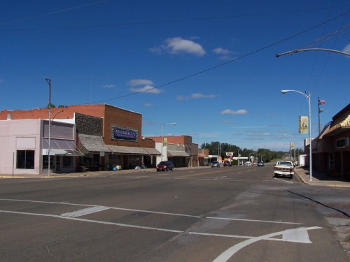 A quiet street in a small town, featuring storefronts, clear blue skies, and a few parked cars.