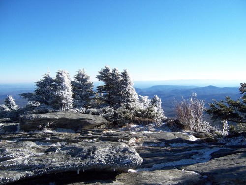 Frost-covered trees on a rocky mountain peak with a clear blue sky and distant hills in the background.