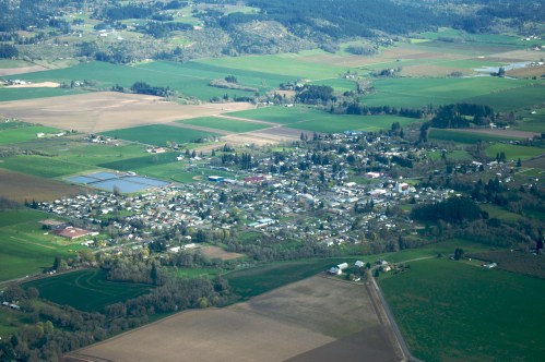 Aerial view of a small town surrounded by green fields and farmland, with scattered buildings and roads.