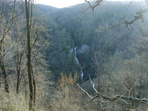 A distant view of a waterfall cascading down a forested hillside, surrounded by bare trees and mountains.
