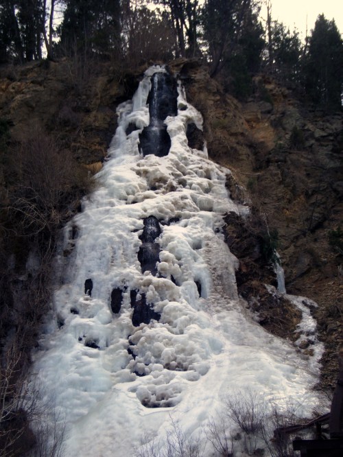 A frozen waterfall cascading down a rocky cliff, surrounded by trees and icy terrain.