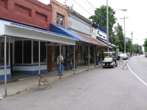 A small town street with brick buildings, blue awnings, and people gathered outside, along with parked vehicles.