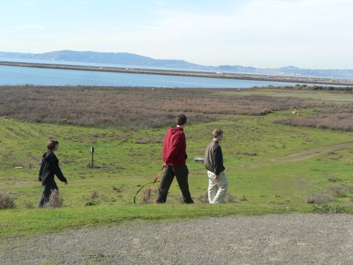 Three people walking along a grassy path near a body of water, with hills and a clear sky in the background.