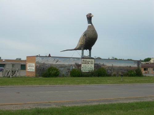 A large pheasant statue stands atop a welcome sign for Huron, SD, with a mural depicting wildlife below.