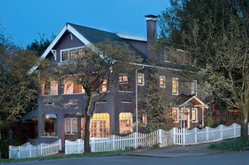 A charming two-story house with a white picket fence, surrounded by trees, illuminated at dusk.