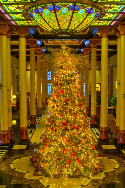 A beautifully decorated Christmas tree with lights and ornaments, set in an elegant lobby with stained glass and columns.