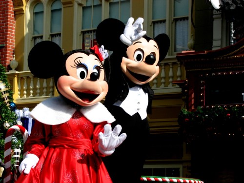 Mickey and Minnie Mouse in festive attire, waving cheerfully at a parade in a colorful setting.