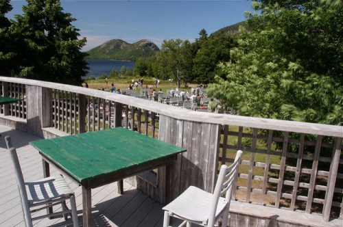 A scenic view from a wooden deck overlooking a lake and mountains, with tables and chairs in the foreground.