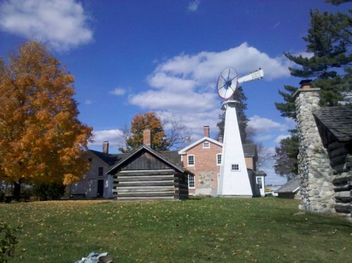 A white windmill stands beside a historic farmhouse and log cabin, surrounded by colorful autumn trees and a blue sky.