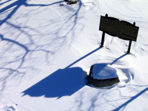 A historical marker partially buried in snow, casting a long shadow on the white surface.