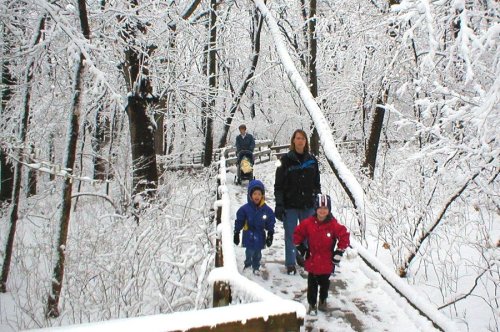 A family walks along a snowy path in a winter forest, surrounded by snow-covered trees.