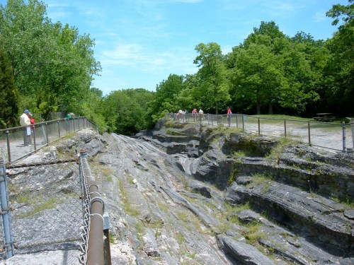 A rocky ravine surrounded by greenery, with a fence and visitors along the path above. Clear blue sky overhead.