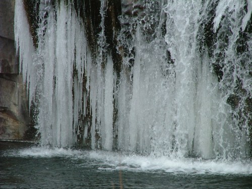 A close-up of a waterfall with icicles hanging from the rocks, cascading water creating a misty spray below.