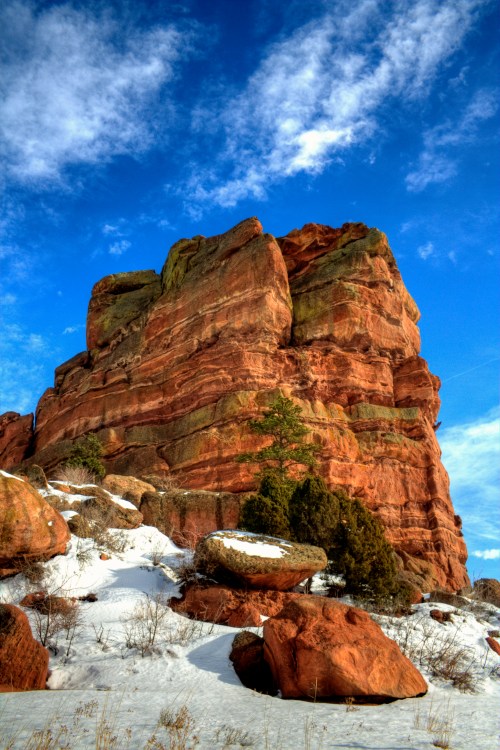 A towering red rock formation against a blue sky, with patches of snow and sparse vegetation at its base.