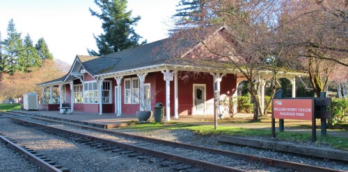Historic red train station building with white trim, surrounded by trees and a sign for William Henry Taylor Railroad Park.