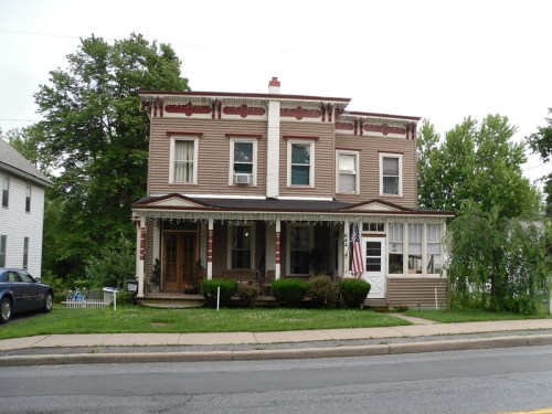 A two-story Victorian-style house with a porch, greenery, and an American flag, set on a quiet street.