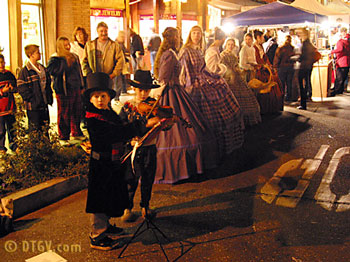 A child plays the violin in front of a crowd, while others in historical costumes stand nearby at a nighttime event.