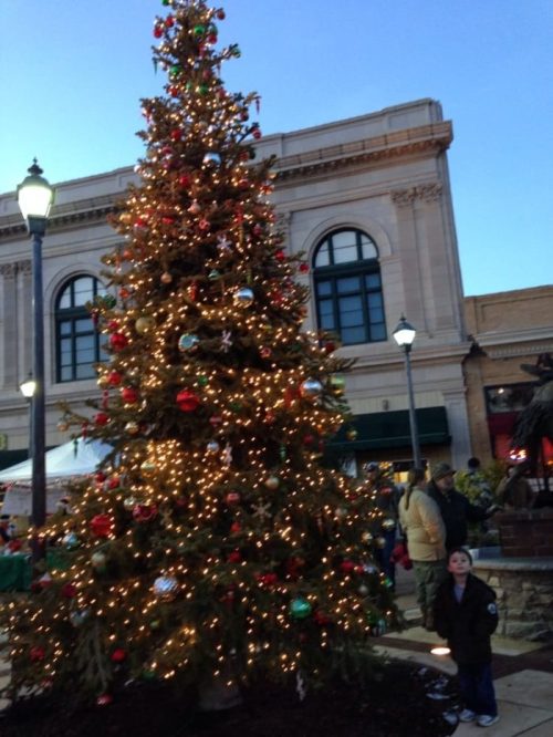 A child stands beside a brightly lit Christmas tree adorned with ornaments, in a festive outdoor setting.