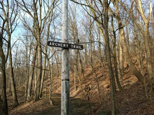 A wooden signpost reading "Archery Trail" in a wooded area with bare trees and a sloped path.