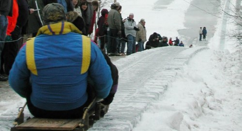 A person in a colorful jacket sleds down a snowy hill, with a crowd of spectators watching from the side.