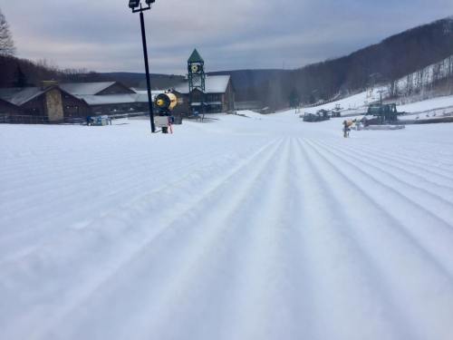 A snowy ski slope with groomed tracks, a lodge in the background, and a cloudy sky overhead.