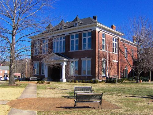 Historic brick building with large windows, a white columned entrance, and benches in a grassy area under a clear blue sky.