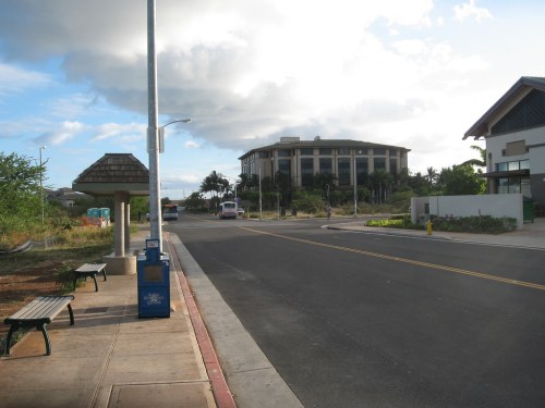 A quiet street scene featuring a bus stop, benches, and a building in the background under a partly cloudy sky.