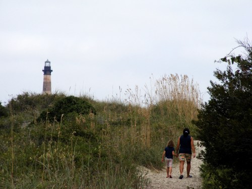 A woman and a child walk along a sandy path, with a lighthouse visible in the background among tall grasses.