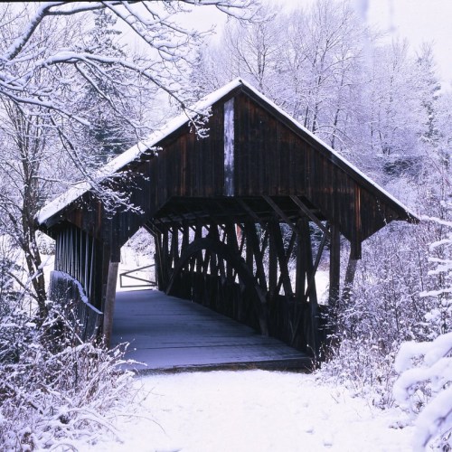 A snow-covered wooden covered bridge surrounded by trees in a winter landscape.