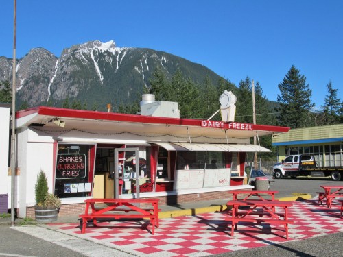 A retro-style diner with red and white decor, picnic tables, and mountains in the background under a clear blue sky.