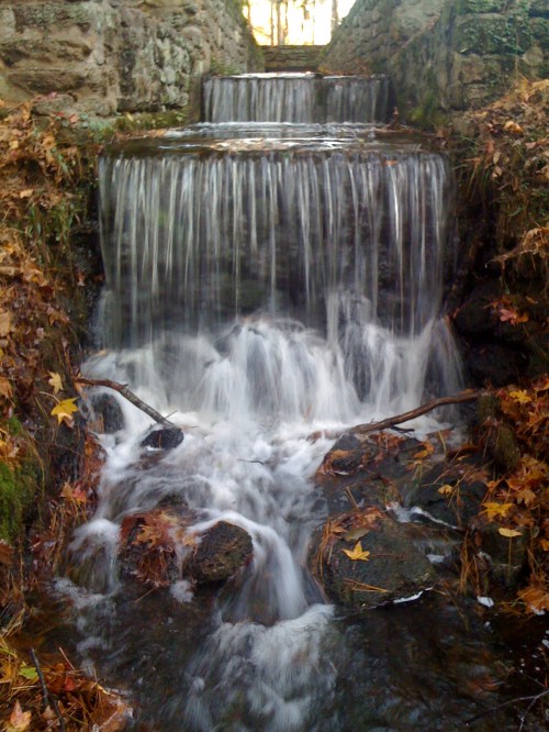 A serene waterfall cascades over rocks, surrounded by autumn leaves and a stone wall in a tranquil setting.