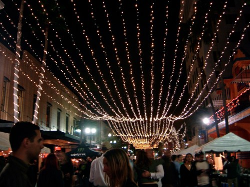 A lively street scene at night, adorned with strings of twinkling lights overhead and people walking below.