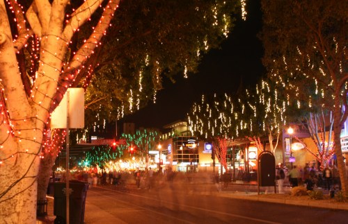 A festive street scene at night, adorned with colorful holiday lights and bustling with people.