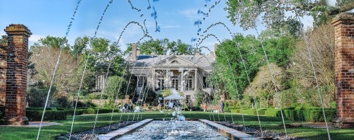 A grand mansion surrounded by lush gardens and fountains, with people enjoying the scenic outdoor space.