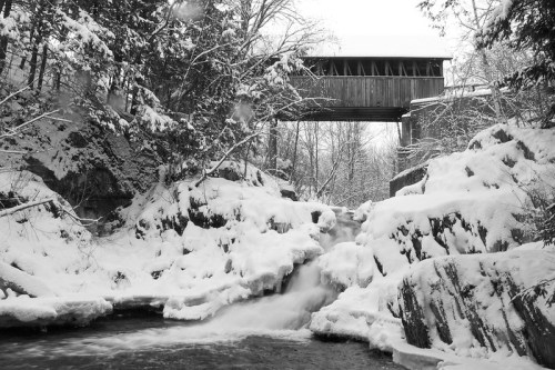 A snowy landscape featuring a covered bridge above a flowing stream, surrounded by rocky terrain and trees.