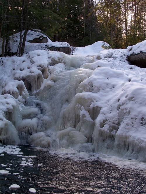 A frozen waterfall cascading over rocks, surrounded by snow and trees in a winter landscape.