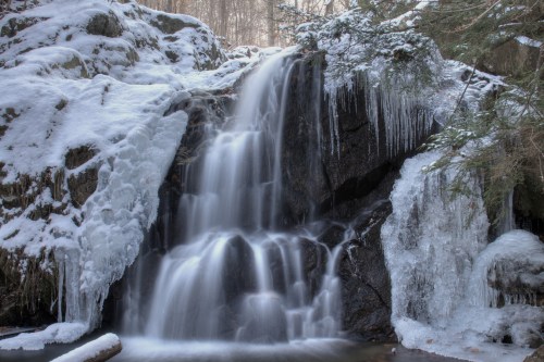 A serene waterfall cascades over rocks, surrounded by snow and icicles in a tranquil winter forest setting.