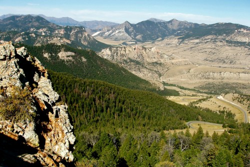 A panoramic view of rugged mountains and valleys, with lush forests and a winding road in the foreground.