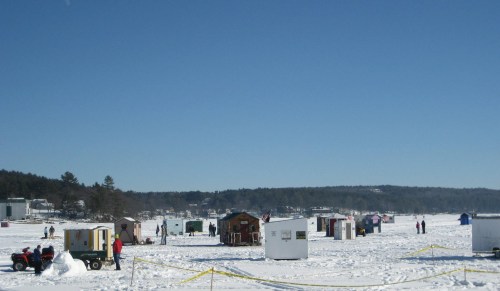 A snowy landscape with several ice fishing huts and people enjoying winter activities under a clear blue sky.