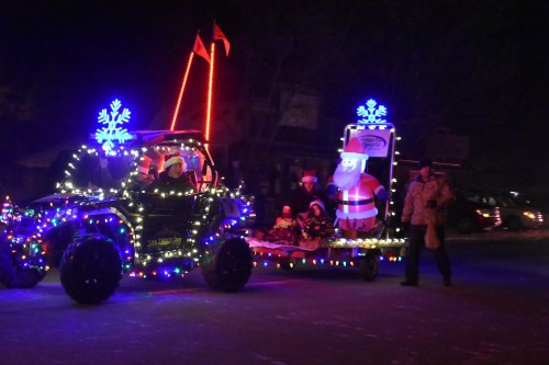 A festive parade scene with a decorated vehicle and a float featuring Santa, surrounded by colorful lights and snowflakes.