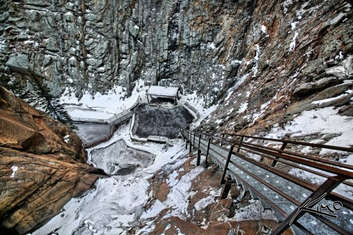 A steep rocky cliff with a metal railing overlooking a snow-covered area and a building below.