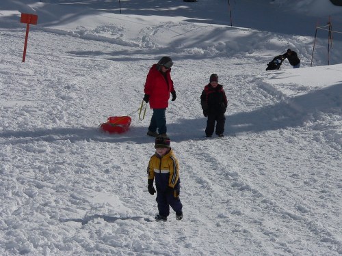 Children playing in the snow, one pulling a sled, while others walk and play in a winter landscape.