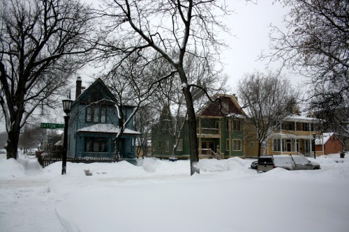 Snow-covered street with historic houses and bare trees on a winter day.