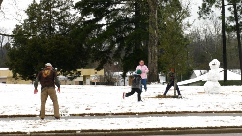 Children playing in the snow, building a snowman, with trees and buildings in the background.