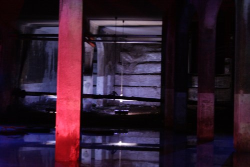 Dimly lit industrial space with reflections on water, featuring red and blue lighting and concrete pillars.