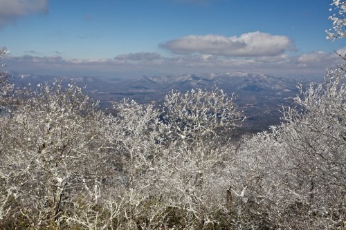 Snow-covered trees in the foreground with a panoramic view of distant mountains under a blue sky with clouds.