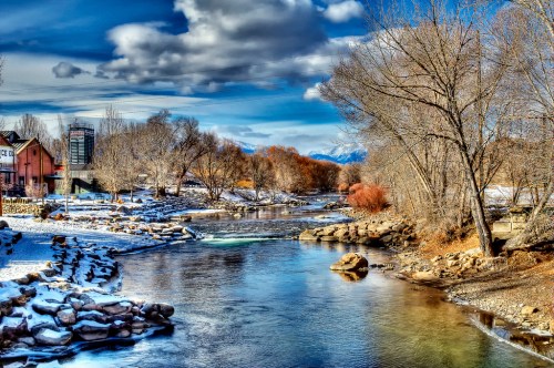A serene river flows through a snowy landscape, surrounded by trees and distant mountains under a cloudy sky.